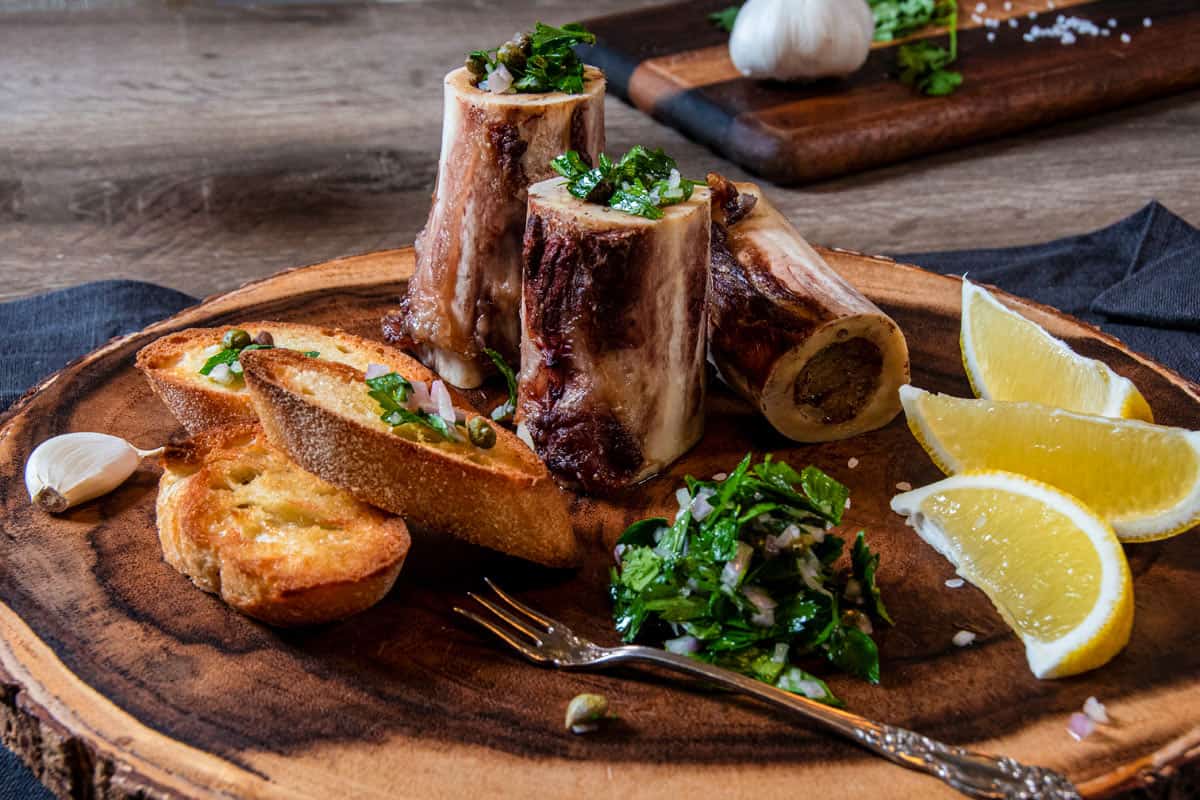 Roasted Marrow Bones with Parsley Salad, Crusted Bread, Lemon Slices, with Coarse Salt, Garlic Bulb and Red Wine Glass
