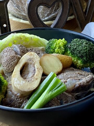beef shank with broccoli, cabbage, green onion and potatoes in a black bowl, with spoon and napkin on the side